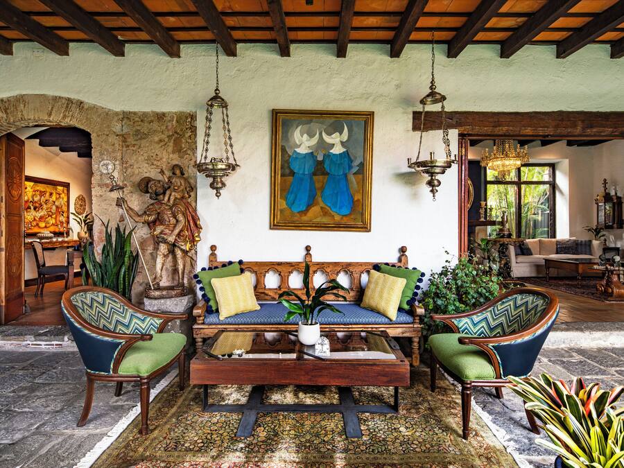 Uma sala de estar acolhedora com um sofá de madeira, duas poltronas e uma mesa de centro. Há uma pintura acima do sofá e uma estátua na parede à esquerda.