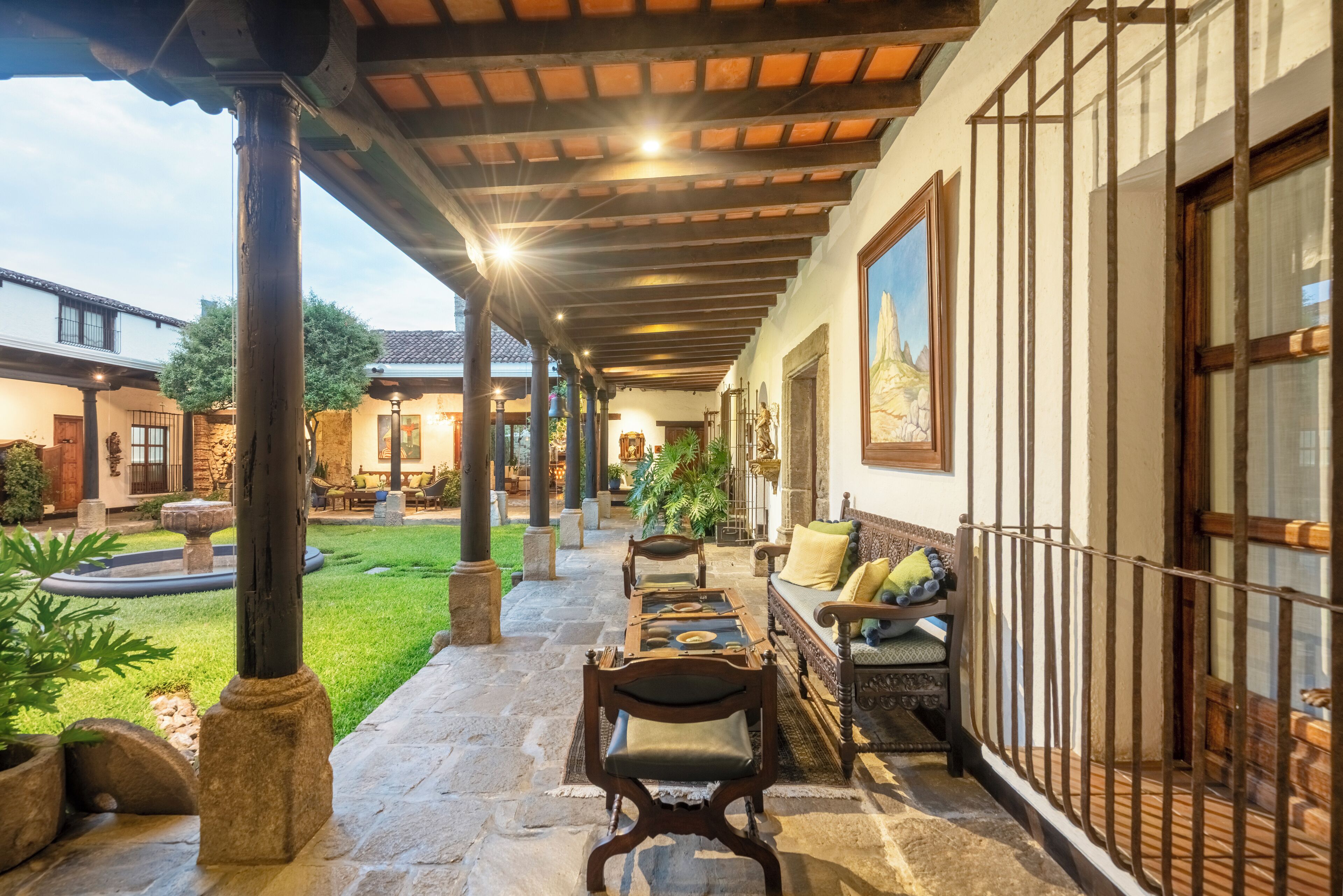 Un porche cubierto con suelo de piedra, mobiliario de madera y vistas a un patio con vegetación y una fuente.