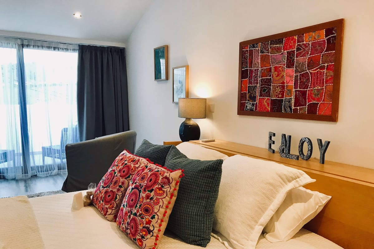 A hotel room with a bed, a nightstand, a lamp, a window with curtains, and a wall with a colorful artwork.
