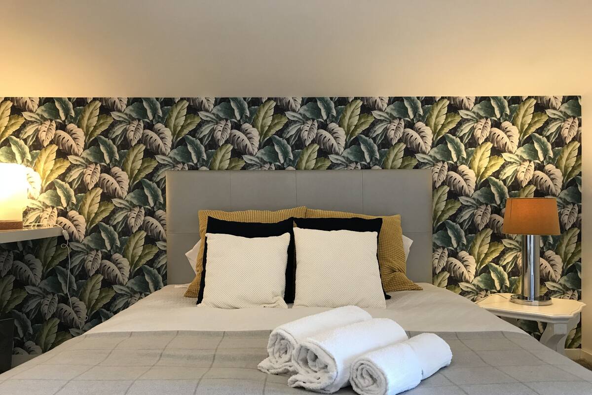 A neatly made bed with a grey quilt, two white pillows, and a rolled white towel.