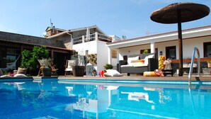 Una piscina cristalina con sillas de descanso y sombrillas, rodeada por un edificio de fachada de piedra y exterior blanco.