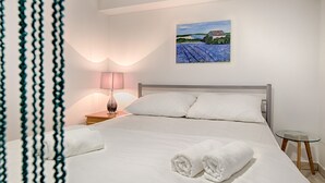 A neatly made bed with white linens and two rolled towels, a bedside lamp, a small bedside table, and a painting of a landscape on the wall. - Cezar Luxury Apartments (Omis)