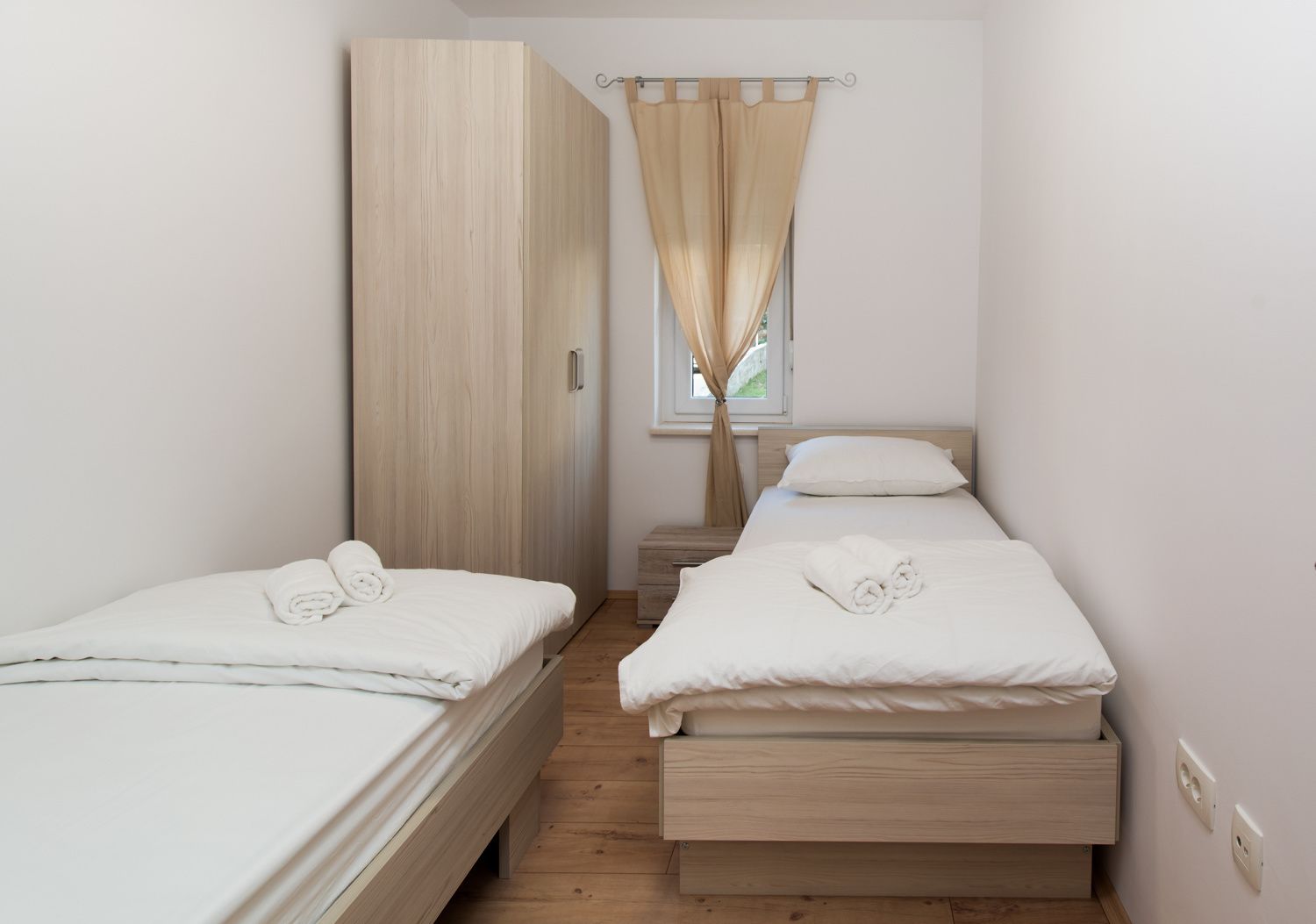 A small, clean room with two single beds, a wooden wardrobe, and a window with curtains.