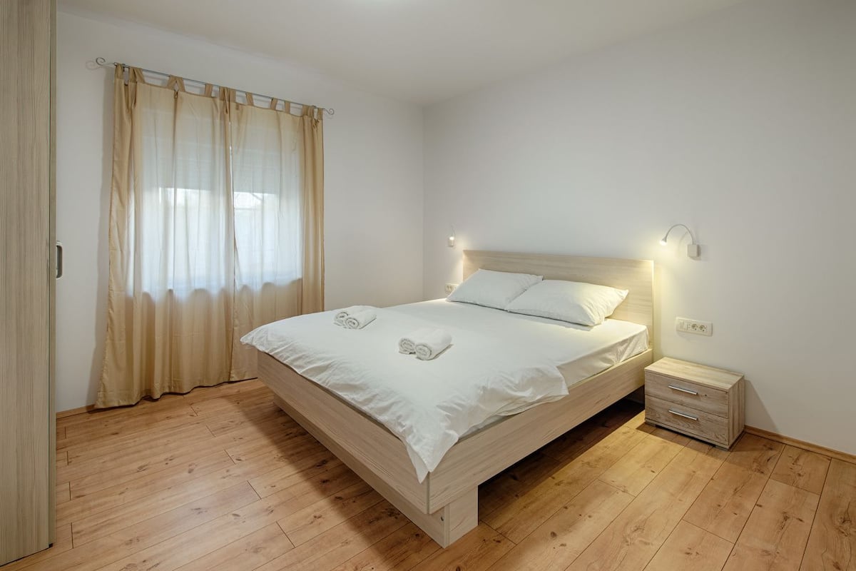 A bedroom with a wooden bed, beige curtains, and a nightstand with a lamp.