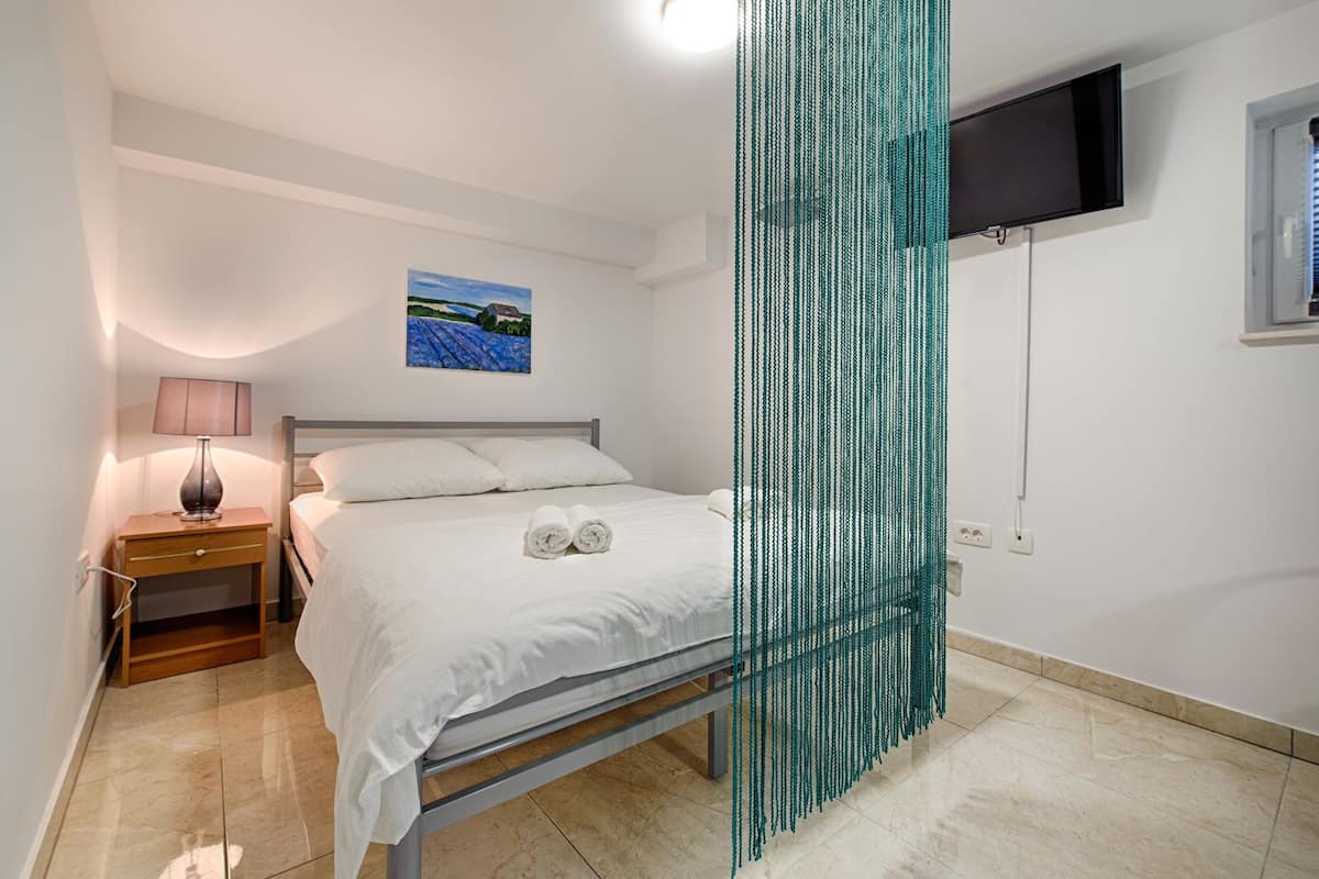 A bedroom with a bed, a nightstand, a lamp, a painting on the wall, and a TV mounted on the wall.