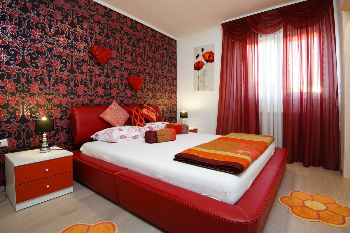 Une chambre avec un lit rouge, une table de chevet avec une lampe, une commode rouge et un papier peint à motifs floraux.