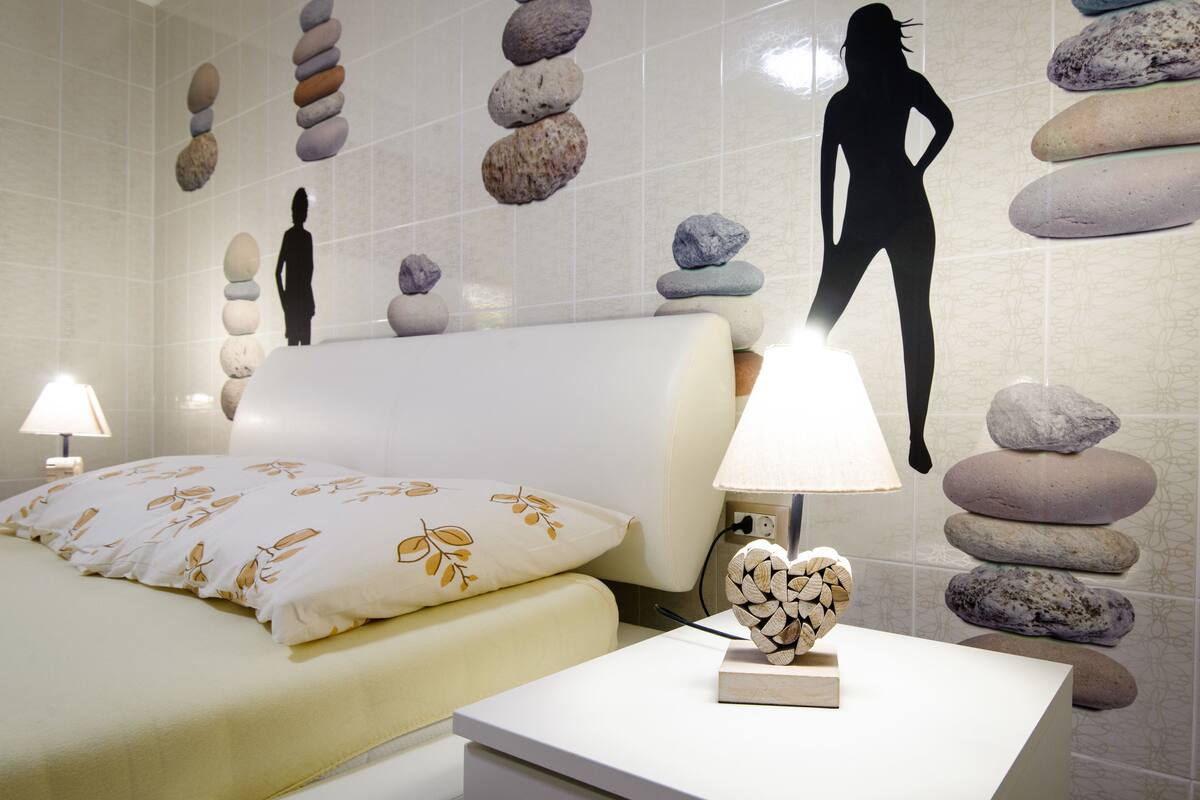 Une chambre d’hôtel avec un lit, une table de chevet avec une lampe et un mur décoré de pierres empilées et de silhouettes de femme.