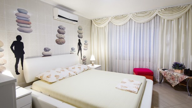 Un dormitorio moderno con una cama grande, una mesita de noche, una silla roja, una pequeña mesa con un florero y una ventana con cortinas.