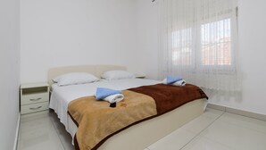 Cama bem arrumada com manta marrom e duas toalhas, mesa de cabeceira branca com gaveta e janela com cortinas leves.
