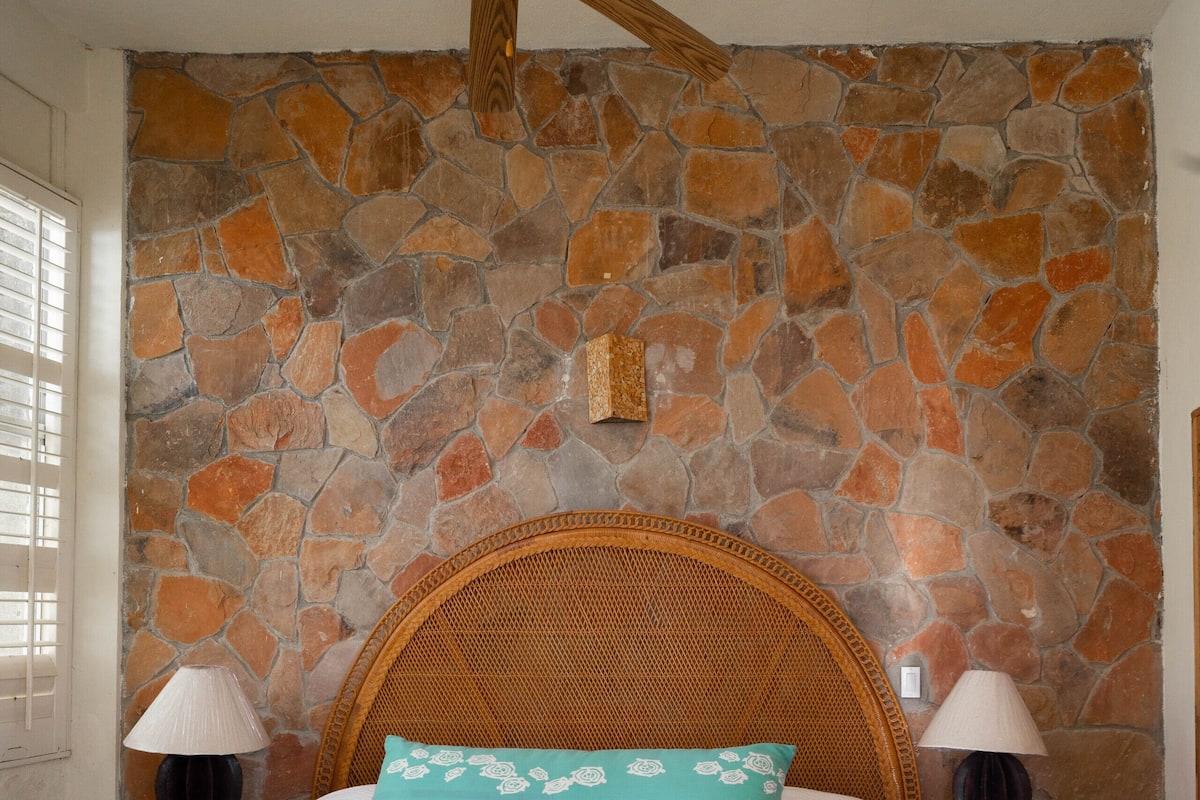 Ein Bett mit Holz-Kopfteil, zwei Nachttische mit Lampen und eine Steinwand.