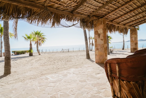 Una playa con una estructura de techo de paja, palmeras y un sendero de piedra que lleva al mar.