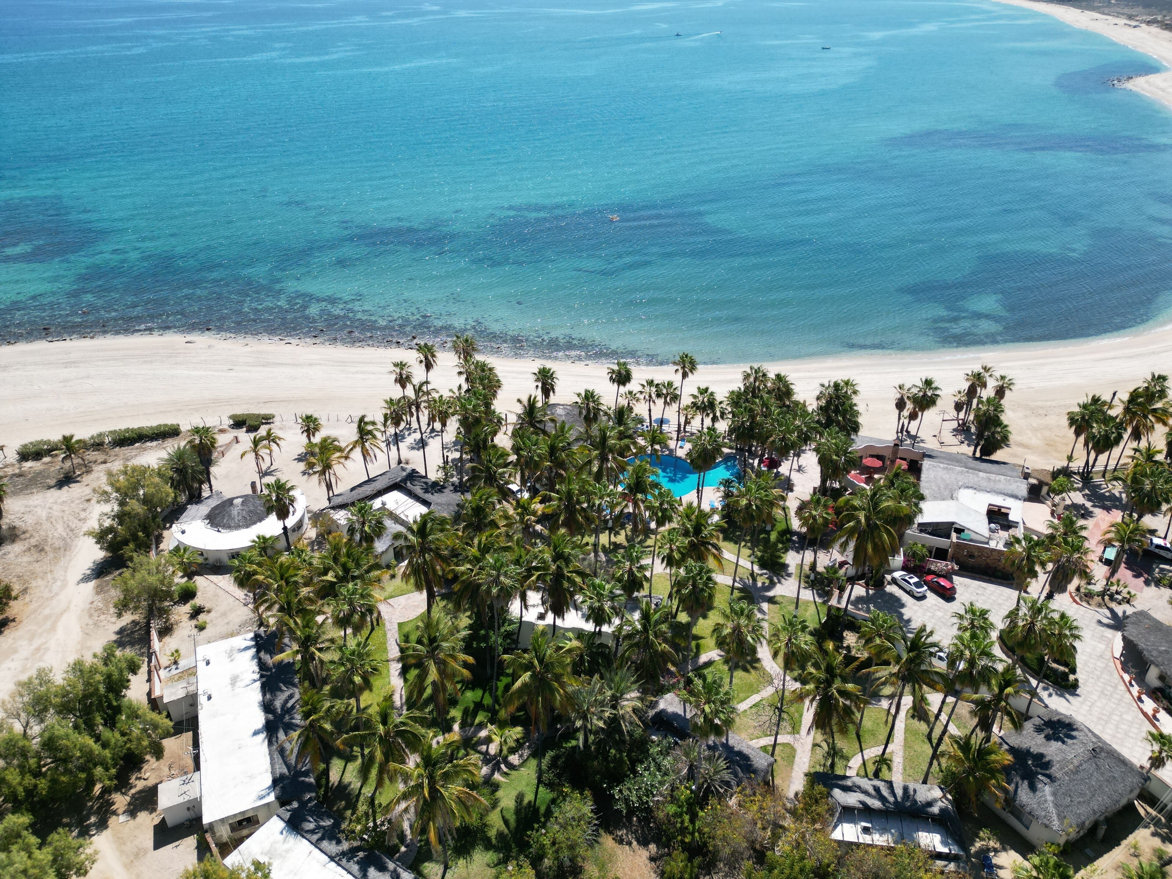 An aerial view of a resort with a swimming pool surrounded by palm trees, a beach, and clear blue waters.