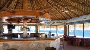 A bar area with a stone counter, wooden stools, and a view of the sea through large windows. - Hotel Rancho Buena Vista (Buenavista)