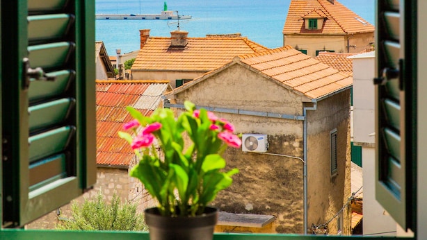 Una ventana con contraventanas abiertas, una maceta con flores rosadas y vista a un pueblo costero con edificios y el mar.