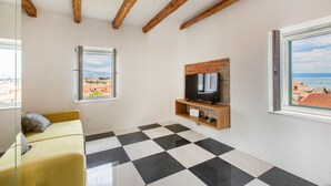 Una sala de estar moderna con piso a cuadros, un sofá amarillo, un televisor montado en una pared de ladrillo y ventanas grandes que ofrecen vistas a la ciudad y al mar.
