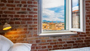 Una habitación con pared de ladrillo, ventana con vista a la ciudad, cama con colcha estampada y una lámpara fijada a la pared.