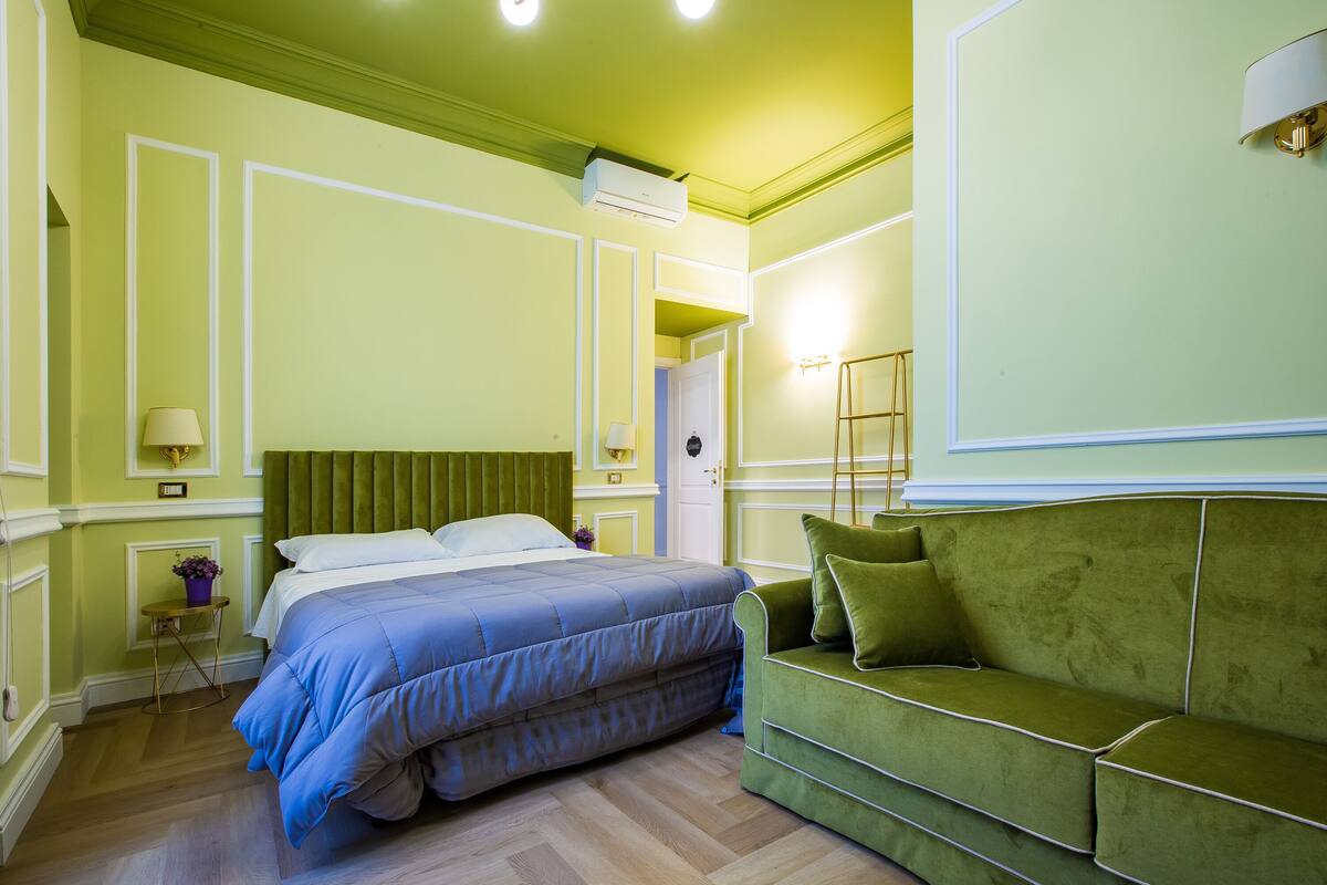 A bedroom with a bed, a green sofa, a bedside table with a lamp, and a door leading to another room.