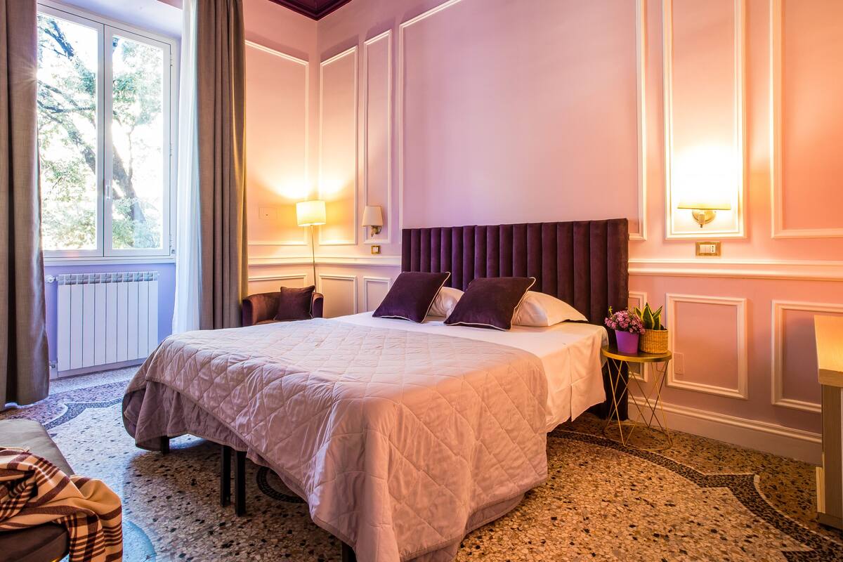 A hotel room with a large bed, two bedside lamps, a small table with a plant, and a window with curtains.