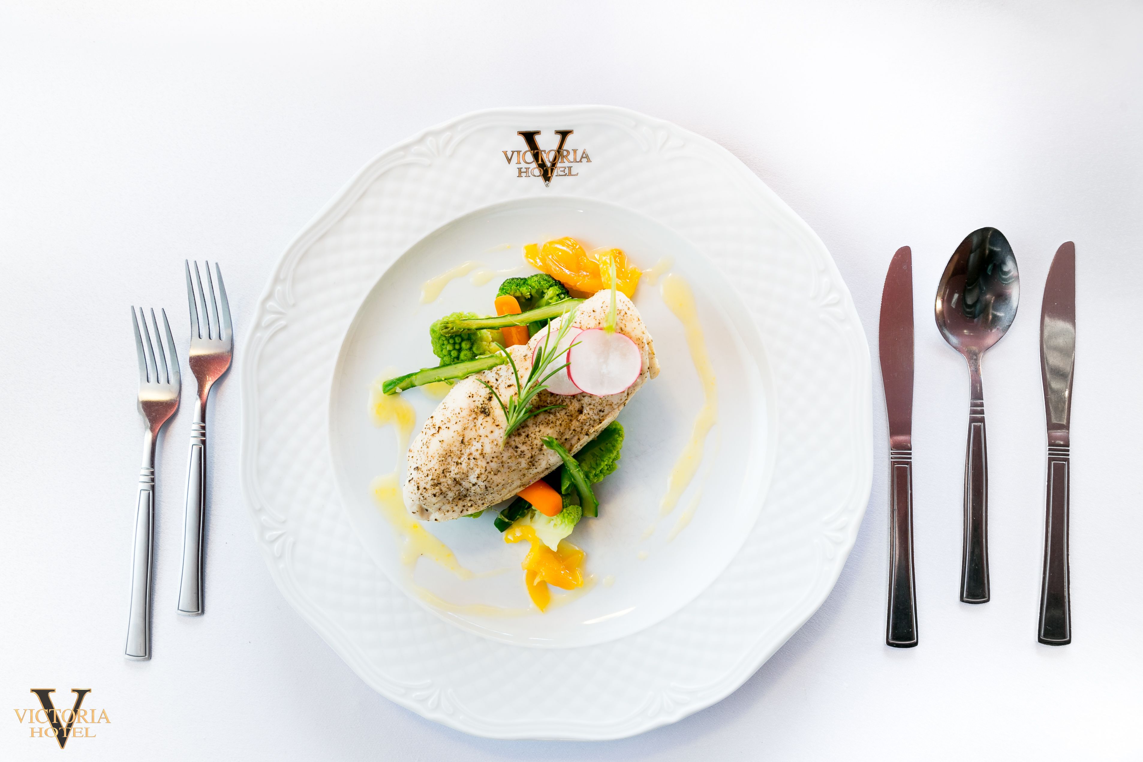 A white plate with a fish dish, vegetables, and sauce, accompanied by cutlery on a white table.