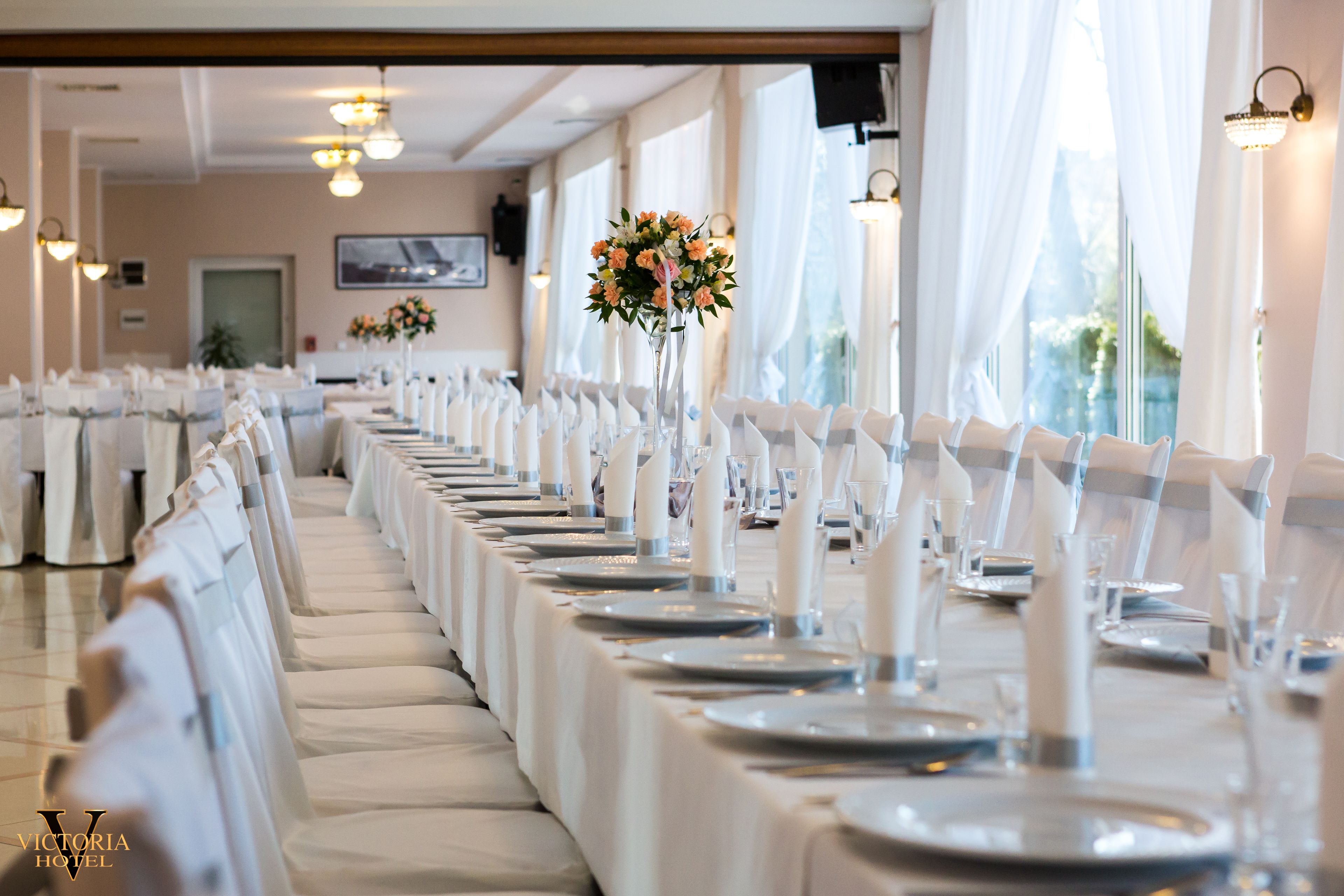 Un salón de banquetes con largas mesas dispuestas para un evento formal, con manteles blancos, copas de cristal y centros de mesa florales.
