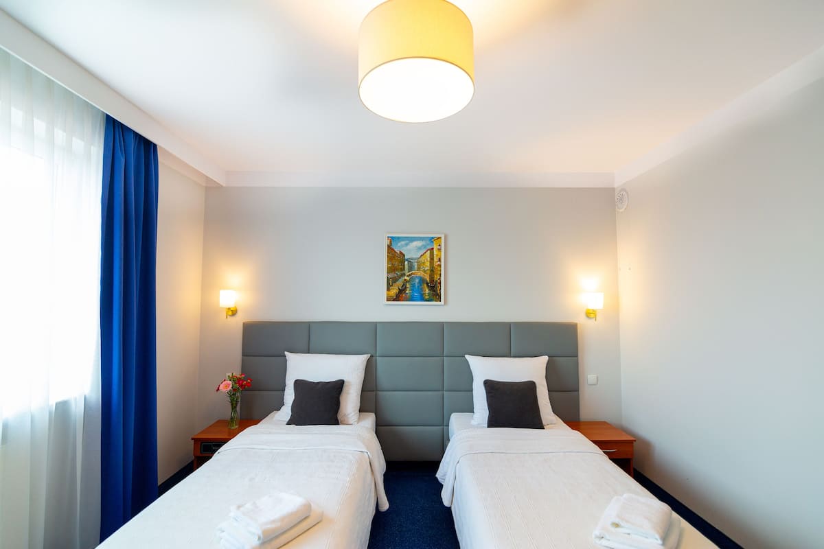 A hotel room with two beds, a painting on the wall, and a window with curtains.