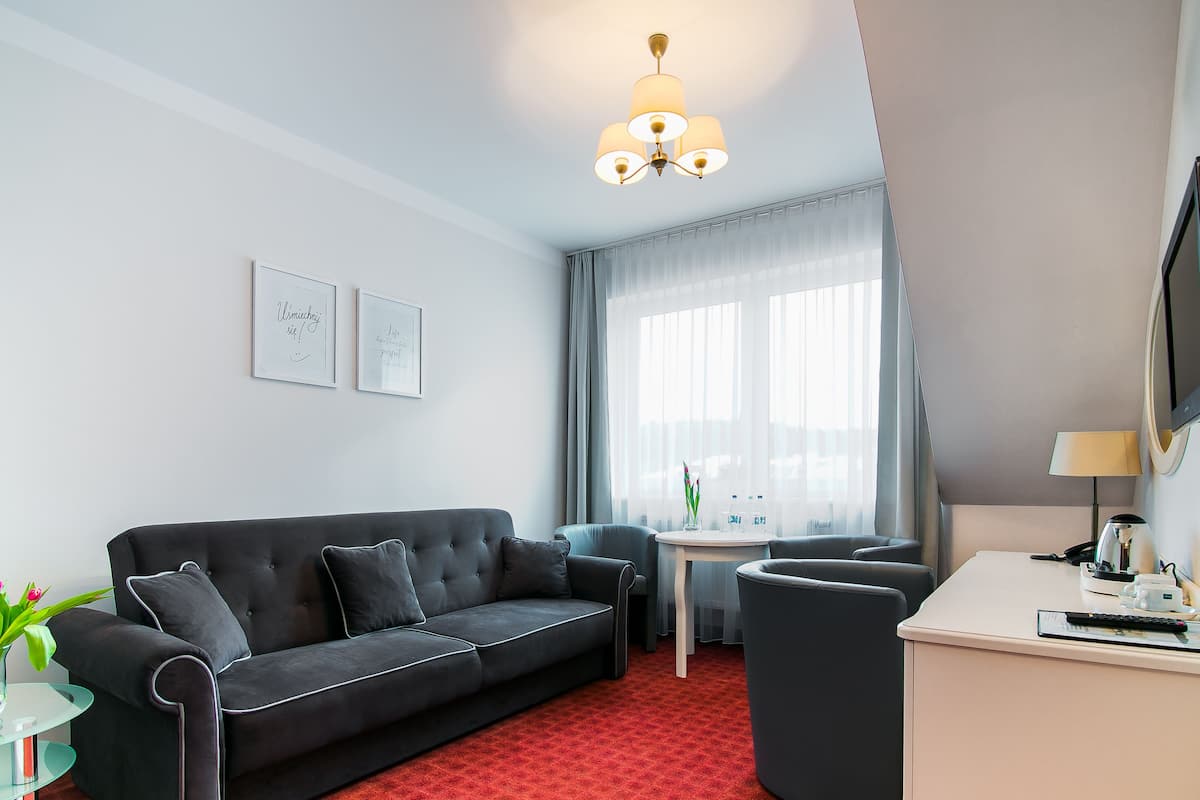A modern hotel room with a black sofa, a small round table, a desk with a computer, and a TV. There are two framed quotes on the wall and a window with sheer curtains.
