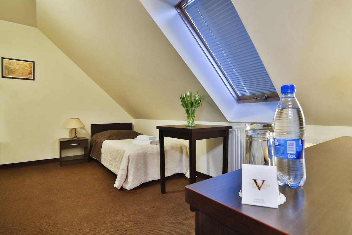 A hotel room with a bed, a small table with a vase, a water bottle, and a sign on the table.