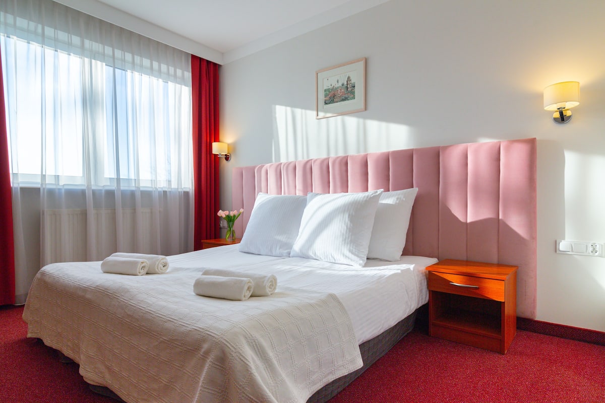 A hotel room with a large bed, a bedside table, a nightstand, a window with curtains, and a wall-mounted lamp.