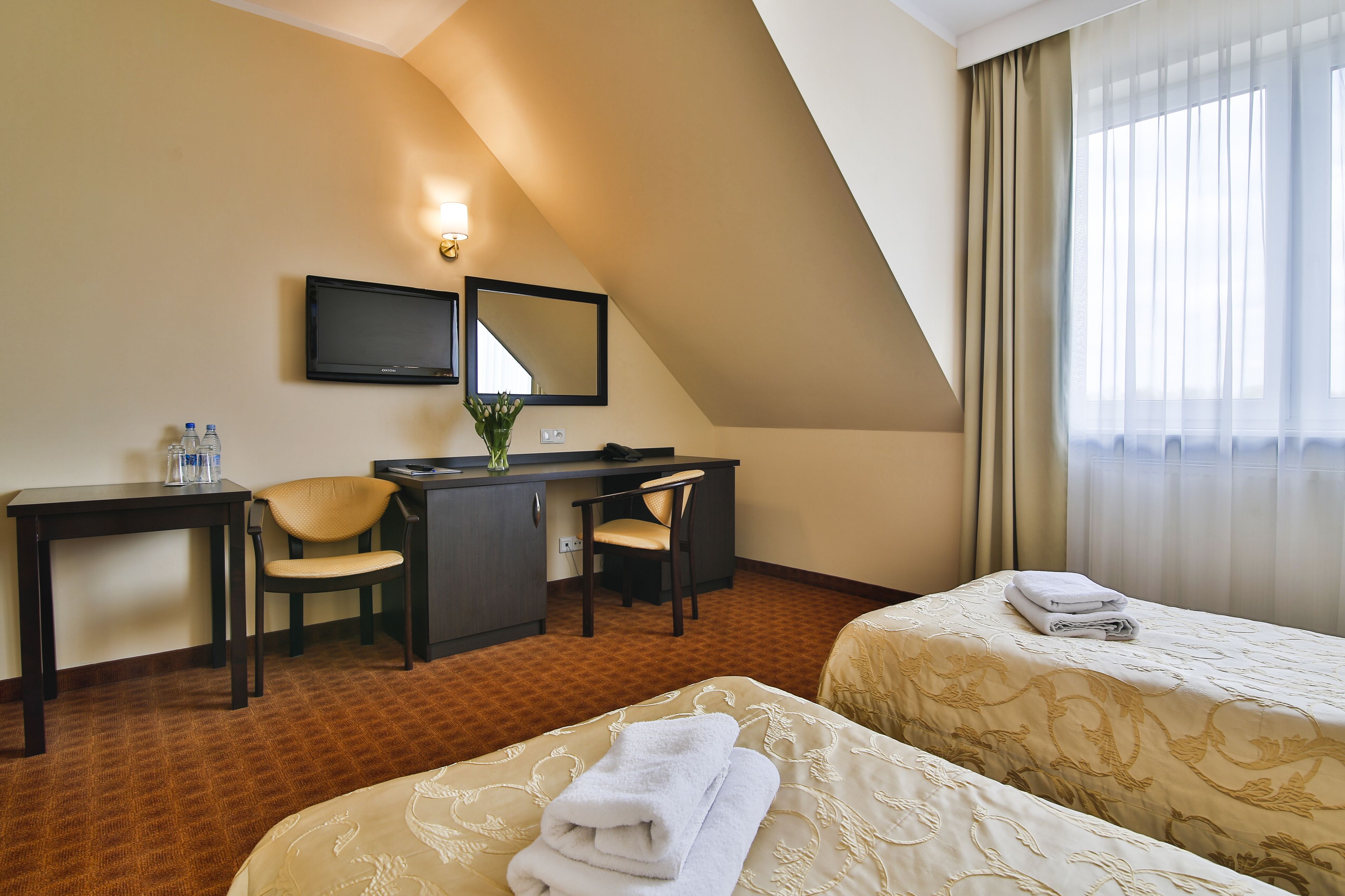 Quarto de hotel com duas camas, uma escrivaninha, uma cadeira, uma televisão e uma janela com cortinas.
