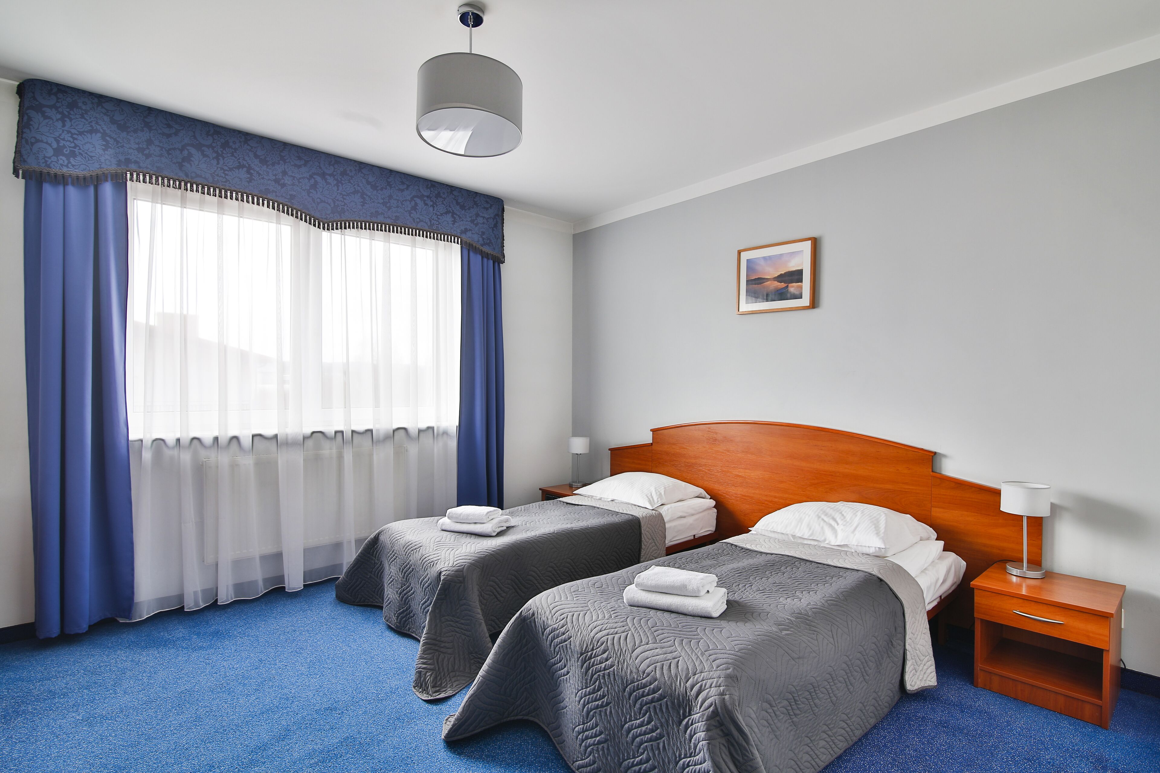 A hotel room with two beds, a wooden headboard, a nightstand with a lamp, and a window with blue curtains.