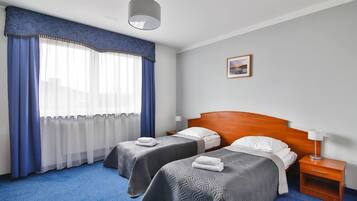 A hotel room with two beds, a wooden headboard, a nightstand with a lamp, and a window with blue curtains.