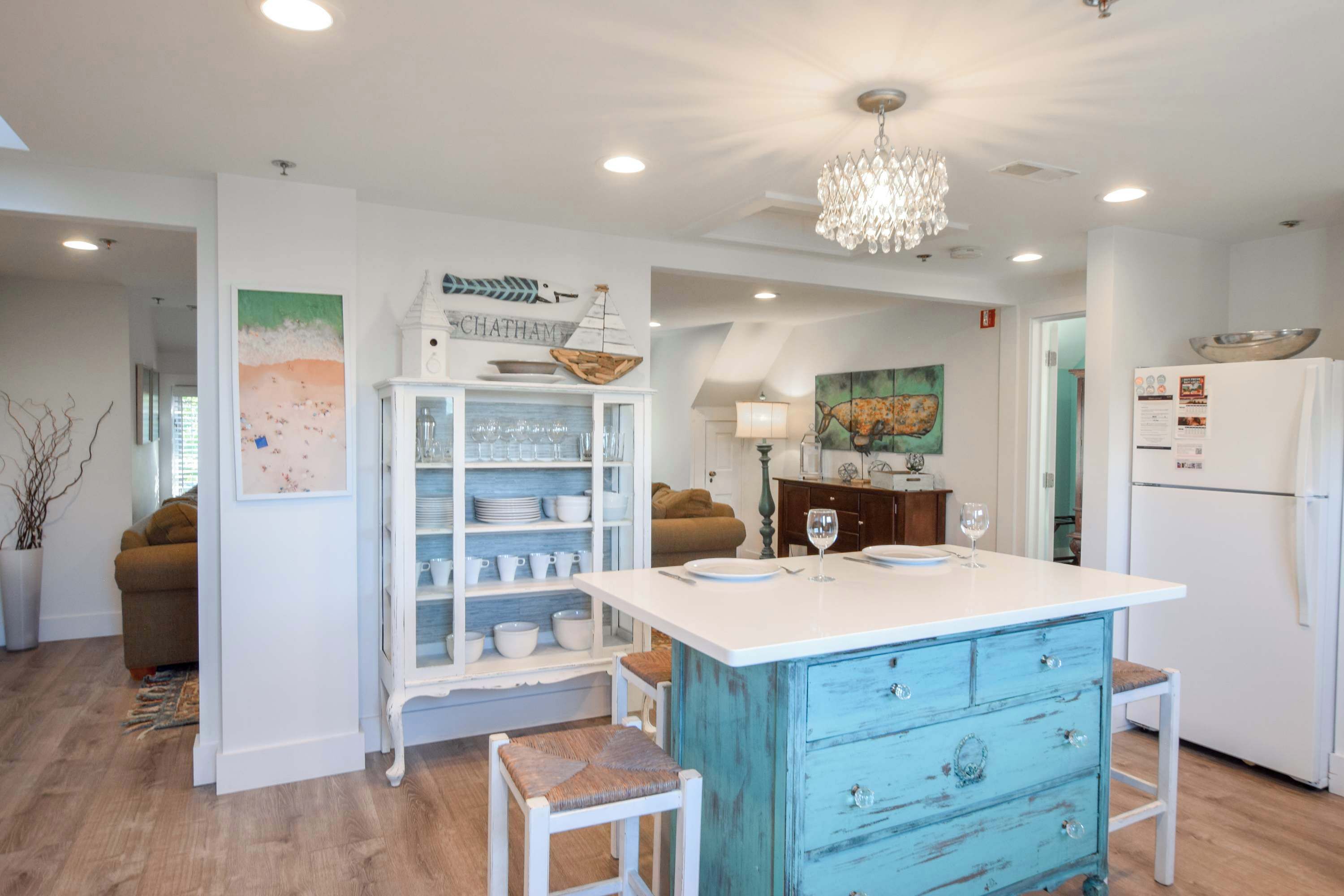 A kitchen with a white island, blue cabinetry, and a chandelier. There are stools and a refrigerator.