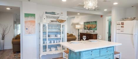 A kitchen with a white island, blue cabinetry, and a chandelier. There are stools and a refrigerator.