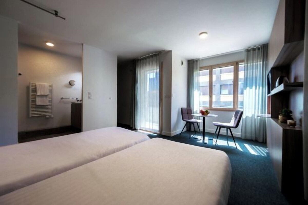 A hotel room with two beds, a small table with chairs, a window with curtains, and a bathroom with a towel rack.