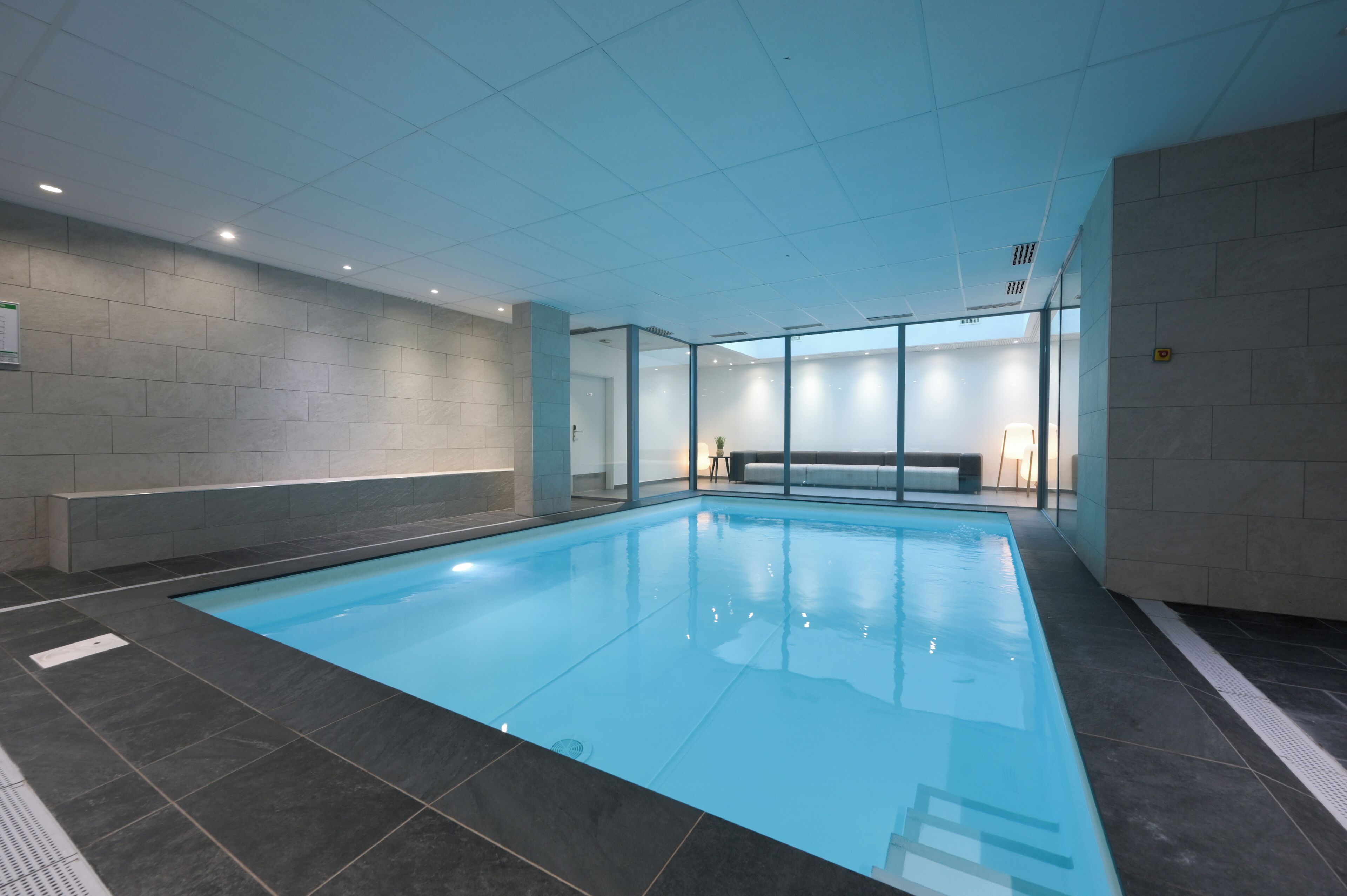 Una piscina interior con suelo y paredes de azulejos, un banco y una pared de vidrio.