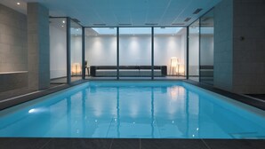 An indoor swimming pool with clear glass walls, a bench, and a ceiling with recessed lighting. - Nemea Appart Hotel So Cloud Lyon Part Dieu (Lyon)