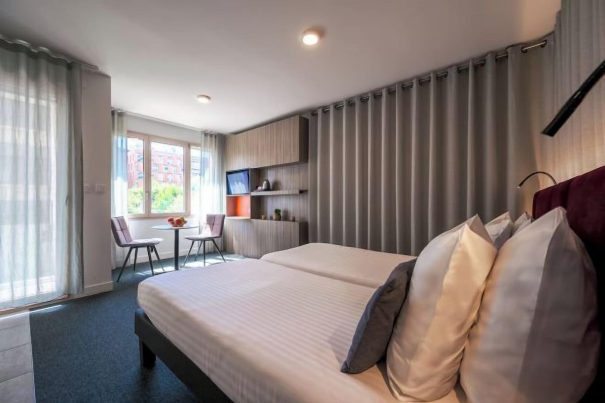 A hotel room with a large bed, a desk, and a chair. There is a window with curtains, a television, and a small table with a vase of flowers.