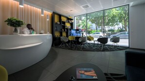A modern reception area with a curved desk, seating with chairs, a large window offering a view of the street, and a bookshelf with various items. - Nemea Appart Hotel So Cloud Lyon Part Dieu (Lyon)