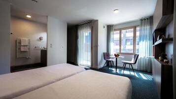 Et hotelværelse med to senge, et lille bord med stole, et vindue med gardiner og et badeværelse med et håndklædetørrestativ.