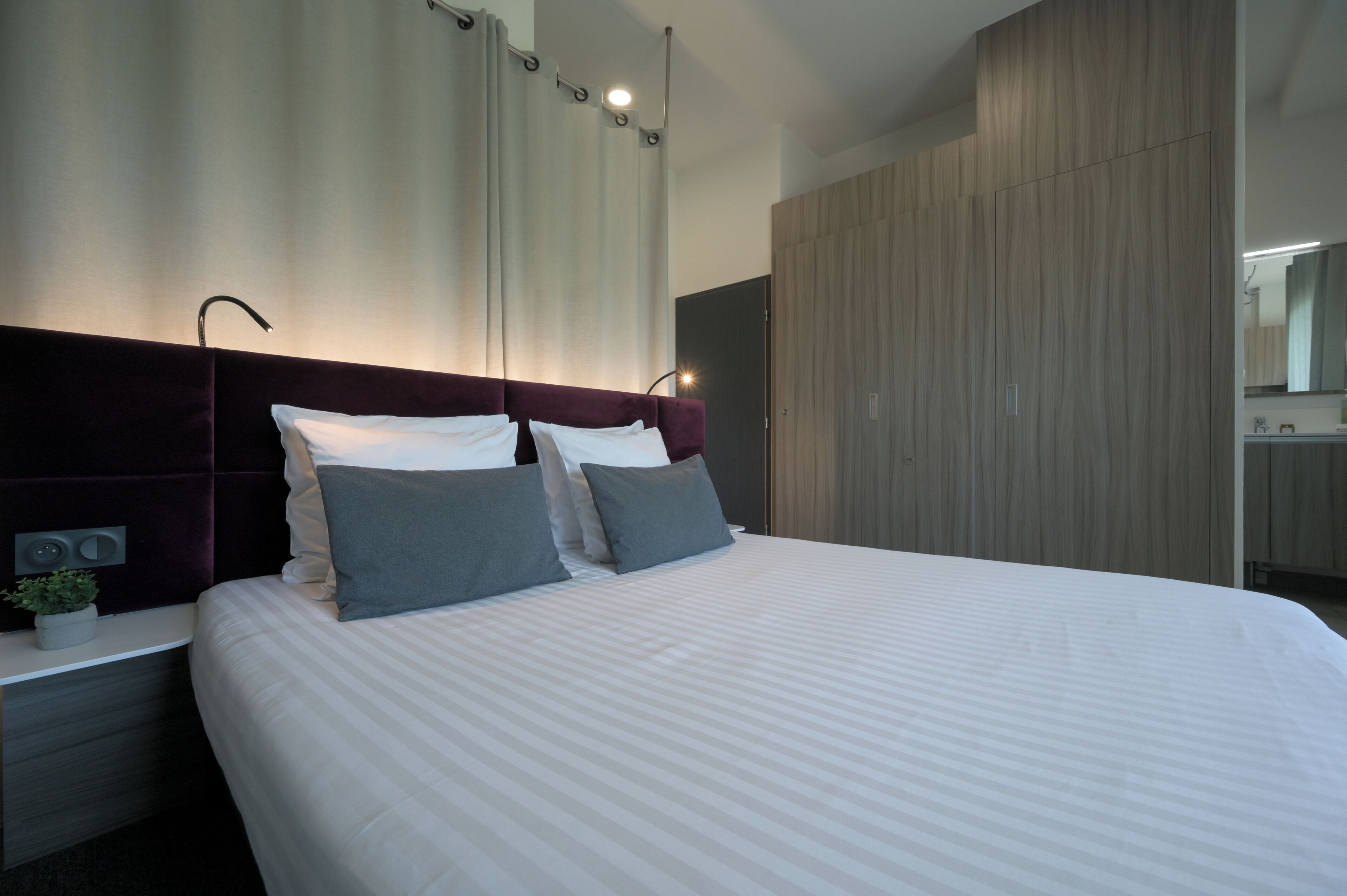Una habitación de hotel moderna con una cama grande, una mesita de noche, una planta en maceta y un armario de madera.