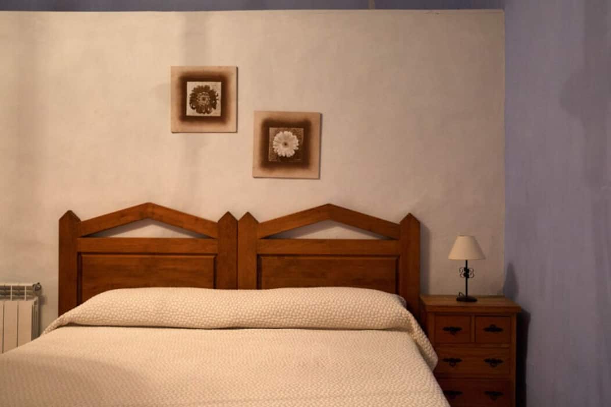 Quarto com cabeceira de madeira, cama com manta branca texturizada, mesa de cabeceira com abajur e duas molduras na parede.