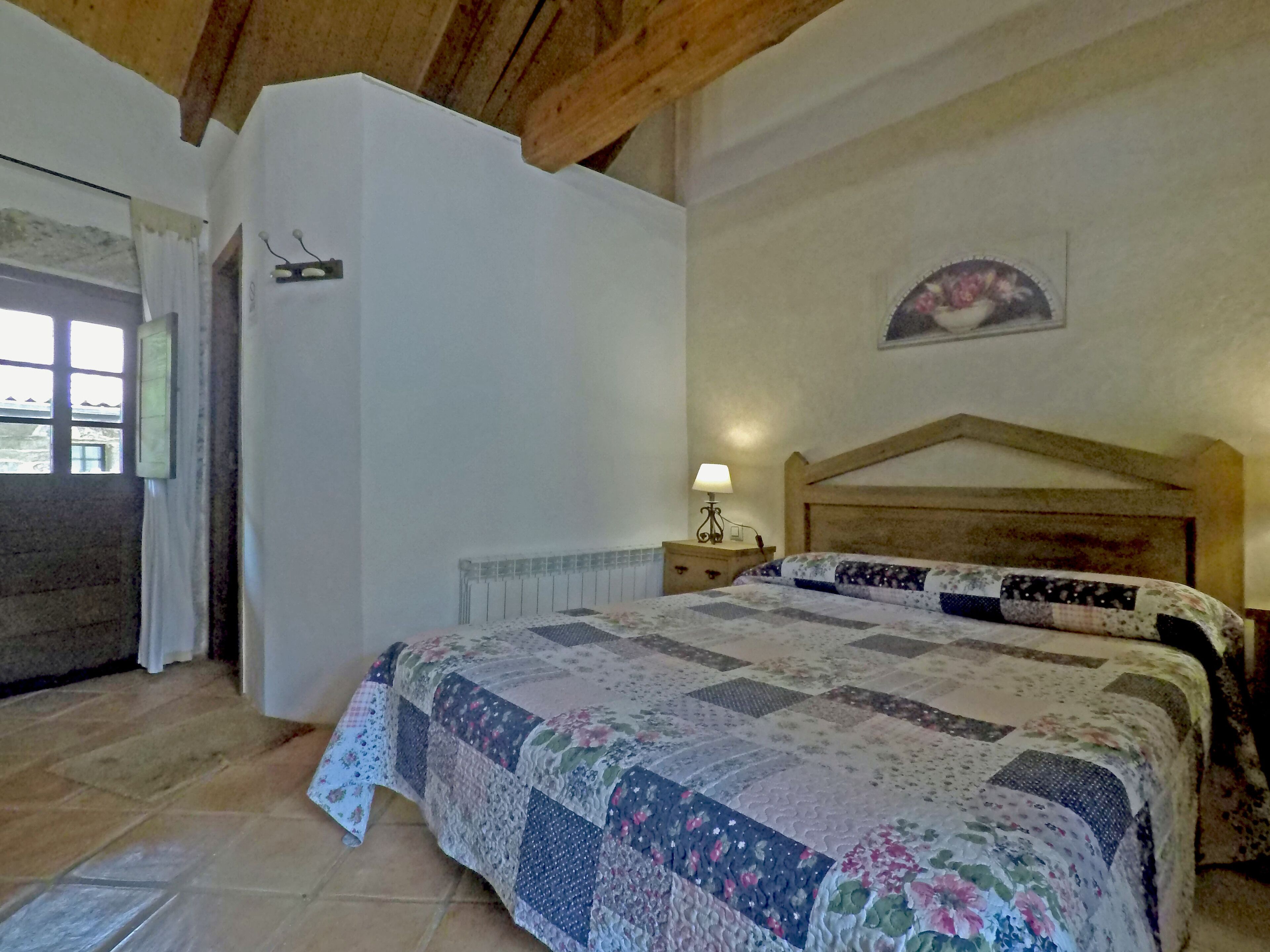 A bedroom with a wooden headboard, a quilted bedspread, a bedside lamp, and a window with curtains.