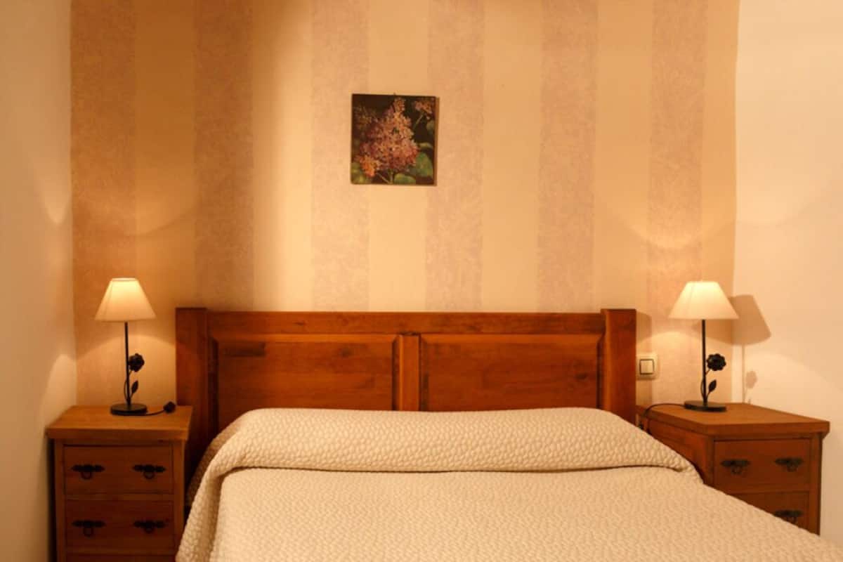 Quarto com cabeceira de madeira, duas mesas de cabeceira com abajures, cama com manta branca texturizada e um painel de parede floral.