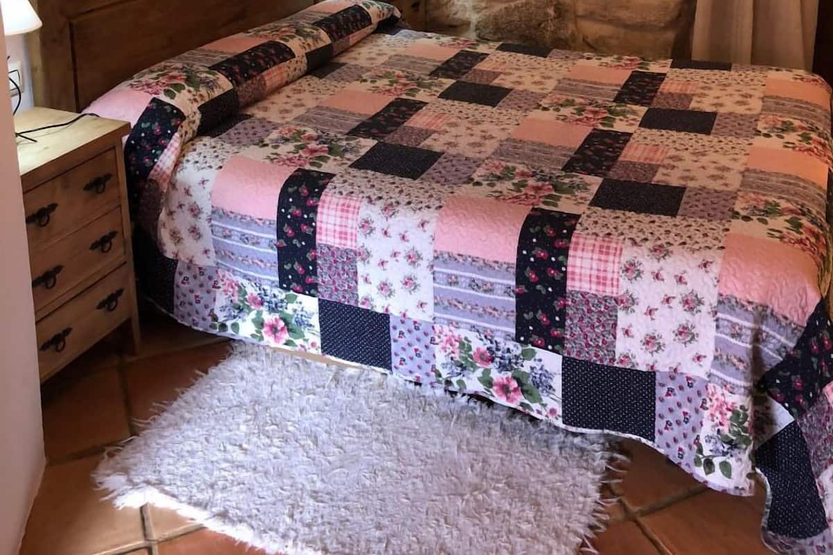 Quarto com parede de pedra, cabeceira de madeira, colcha acolchoada, mesa de cabeceira com luminária e tapete pequeno.