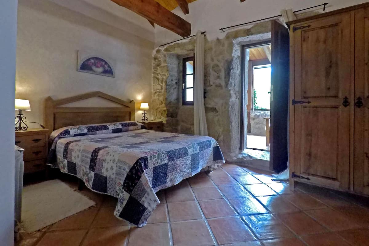 Quarto com cama de madeira, paredes de pedra, guarda-roupa de madeira e janela com vista.