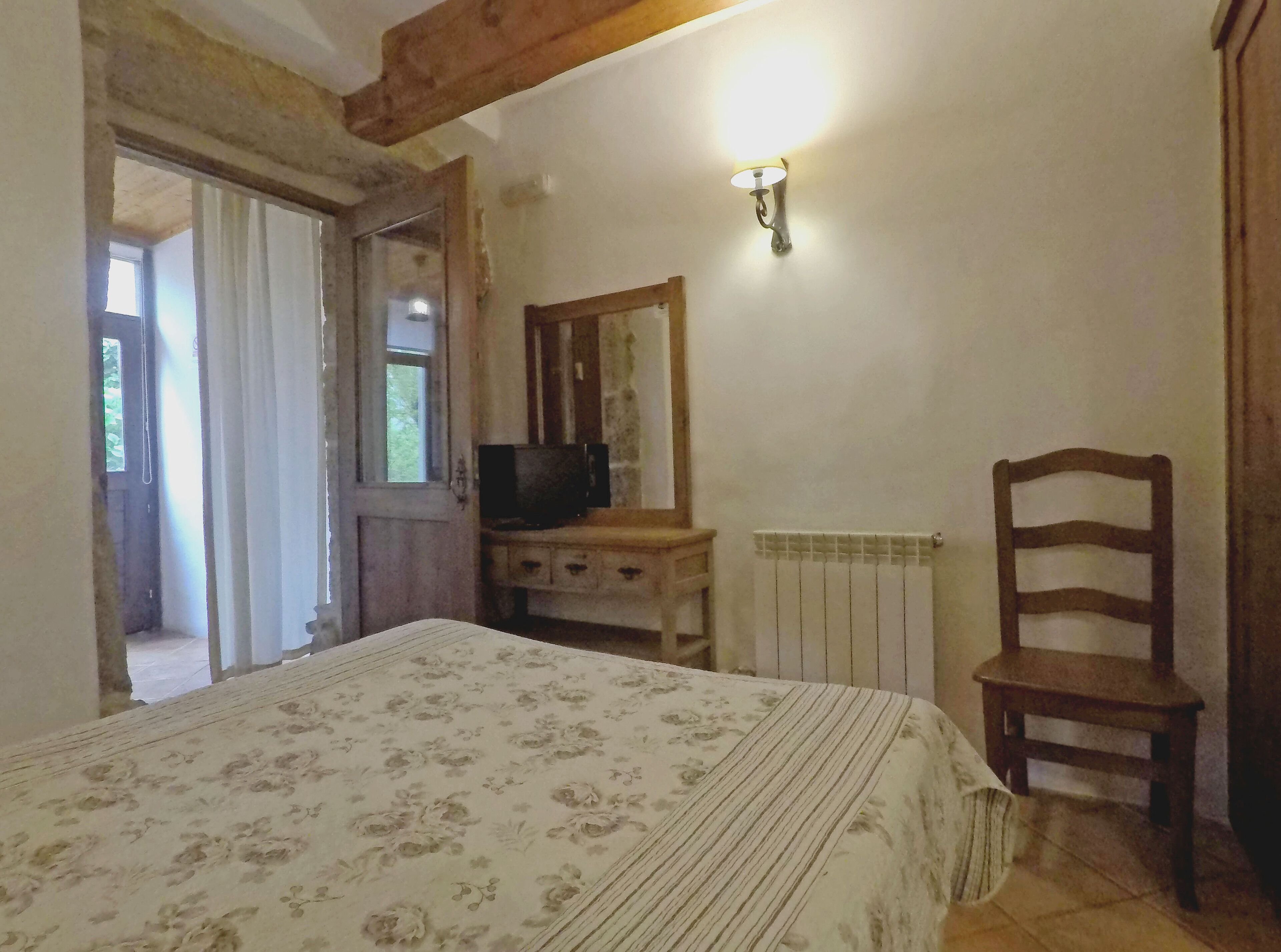Un dormitorio con cama, una silla de madera, cómoda, televisor y lámpara fijada en la pared.