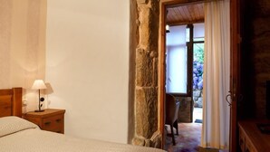 A bedroom with a wooden headboard, a bedside table with a lamp, and a view of an outdoor area with a stone wall and a chair. - Lugar Dos Devas (Covelo)