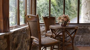 A rustic room with two wicker chairs and a small round table, a vase with flowers, and a view of greenery through large windows. - Lugar Dos Devas (Covelo)
