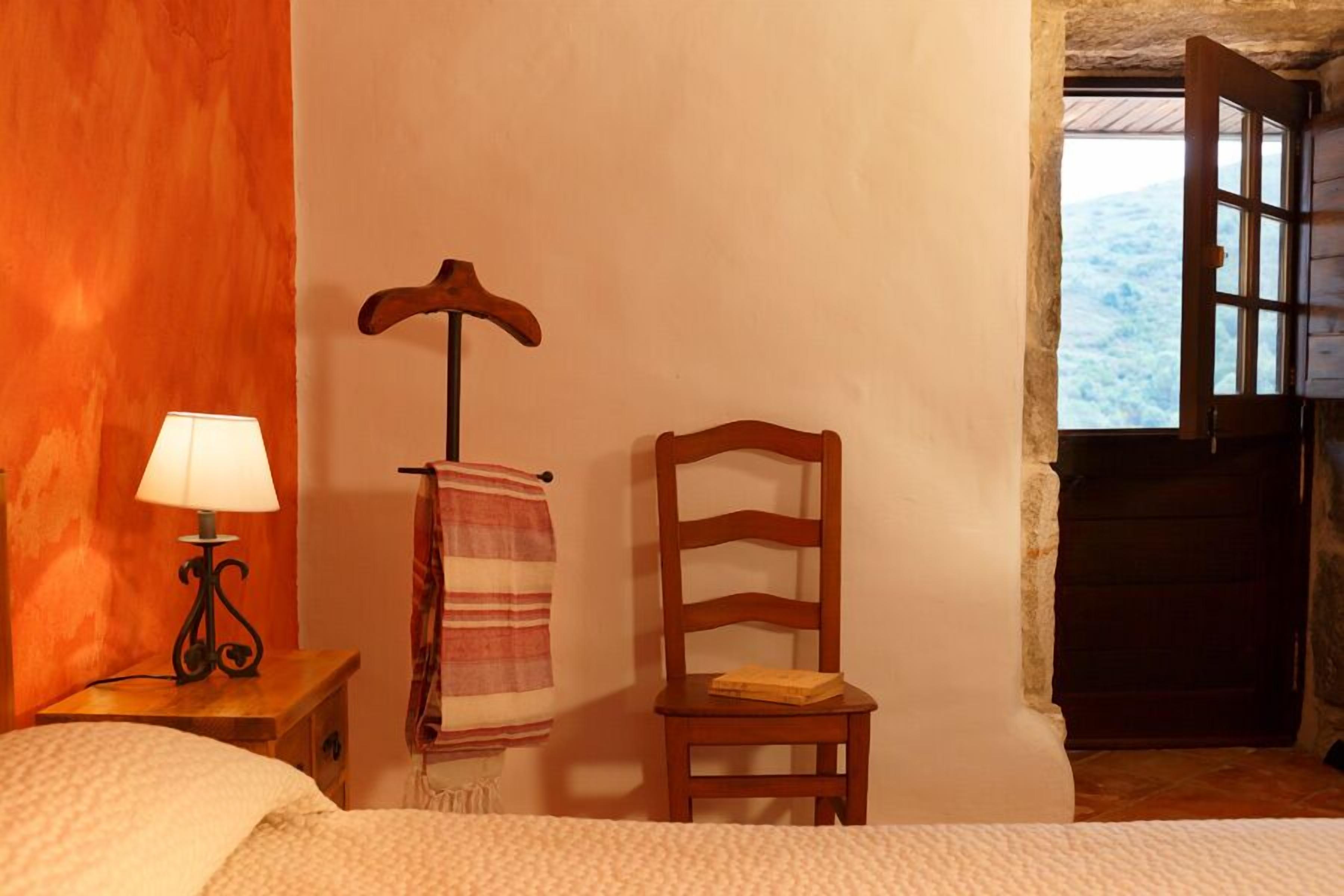 A bedroom with a bed, a nightstand with a lamp, a wooden chair, a towel rack, and a window with a view of the outdoors.