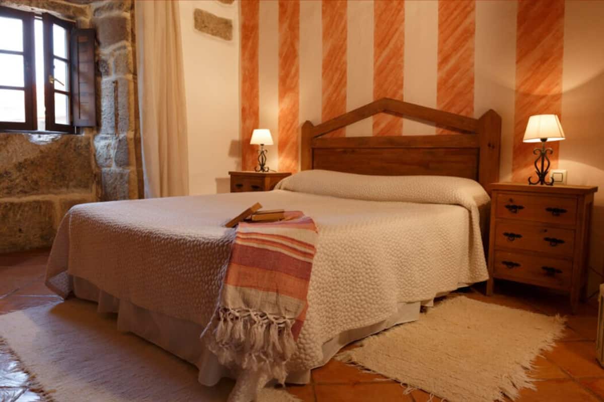 Um quarto aconchegante com uma cama grande, cabeceira de madeira, duas mesas de cabeceira com luminárias, uma parede de pedra e uma janela com persianas.
